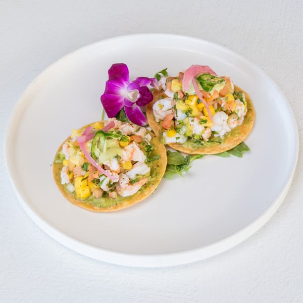 Two shrimp tostadas with avocado, corn, and pickled onions, garnished with a purple orchid.
