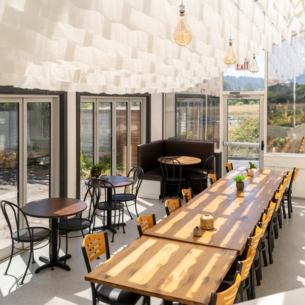 Bright Mi Cava & Cocina dining room with long wooden tables, black chairs, and large windows.