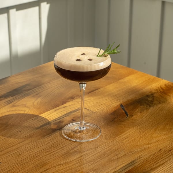 Espresso martini with foam, coffee beans, and a rosemary sprig on a wooden table.